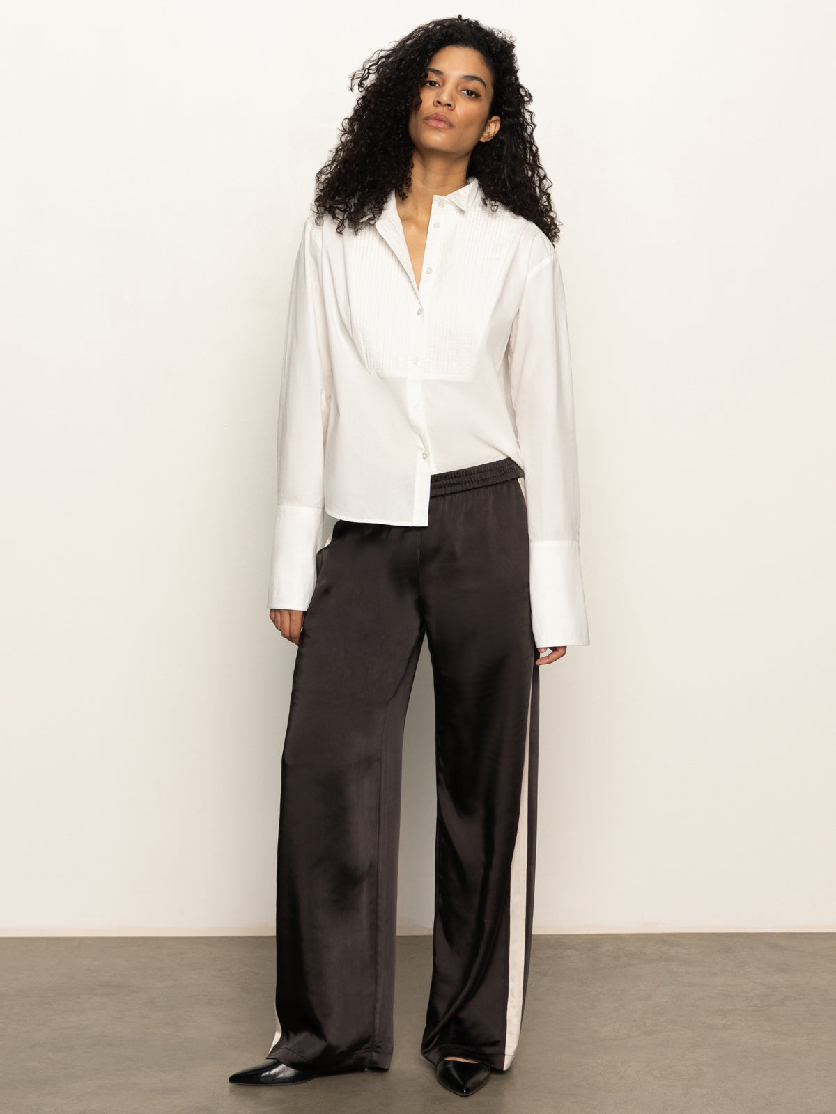 A woman with curly hair stands against a plain background, wearing Sanctuary Clothing's satin track pant in black with white side stripes, paired with a white button-up shirt and black pointed shoes.