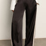 Someone wears the Sanctuary Clothing satin track pant in black, featuring white side panels, paired with a white button-down shirt and black pointed-toe shoes, standing on a gray floor against a plain wall.