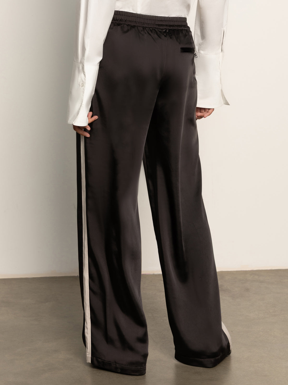 A person wearing Sanctuary Clothing satin track pant black, featuring white side stripes, and a white long-sleeve shirt stands on a concrete floor facing away from the camera.