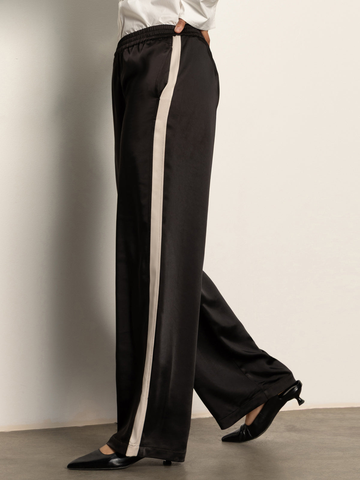 A person models the Sanctuary Clothing satin track pant in black, featuring a light beige side stripe, styled with black low-heeled shoes and a white top against a plain light wall.