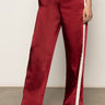 A person wears Sanctuary Clothing's satin track pant crimson€�shiny red with white side stripes and a relaxed fit€�paired with red heeled shoes. The photo, cropped at the torso, highlights the standout pants.
