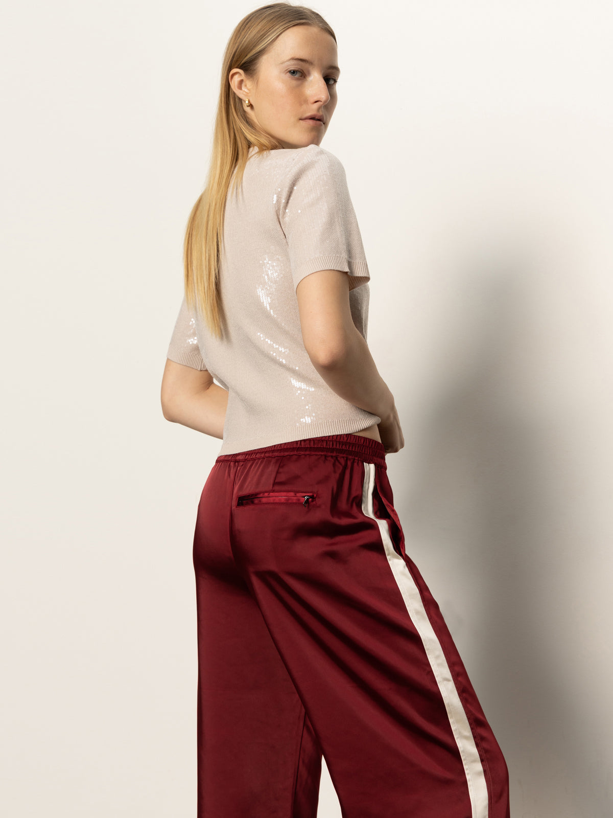 A woman with long blonde hair models the Sanctuary Clothing satin track pant in crimson, featuring white side stripes, paired with a shiny beige short-sleeve top. She stands against a plain light background, looking over her shoulder.