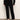 A person stands against a white wall wearing Sanctuary Clothing's 90s straight legging black pants, a black long-sleeve sweater, and black flats. Their face is not visible in the image.