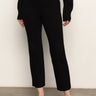 A person stands against a white wall wearing Sanctuary Clothing's 90s straight legging black pants, a black long-sleeve sweater, and black flats. Their face is not visible in the image.