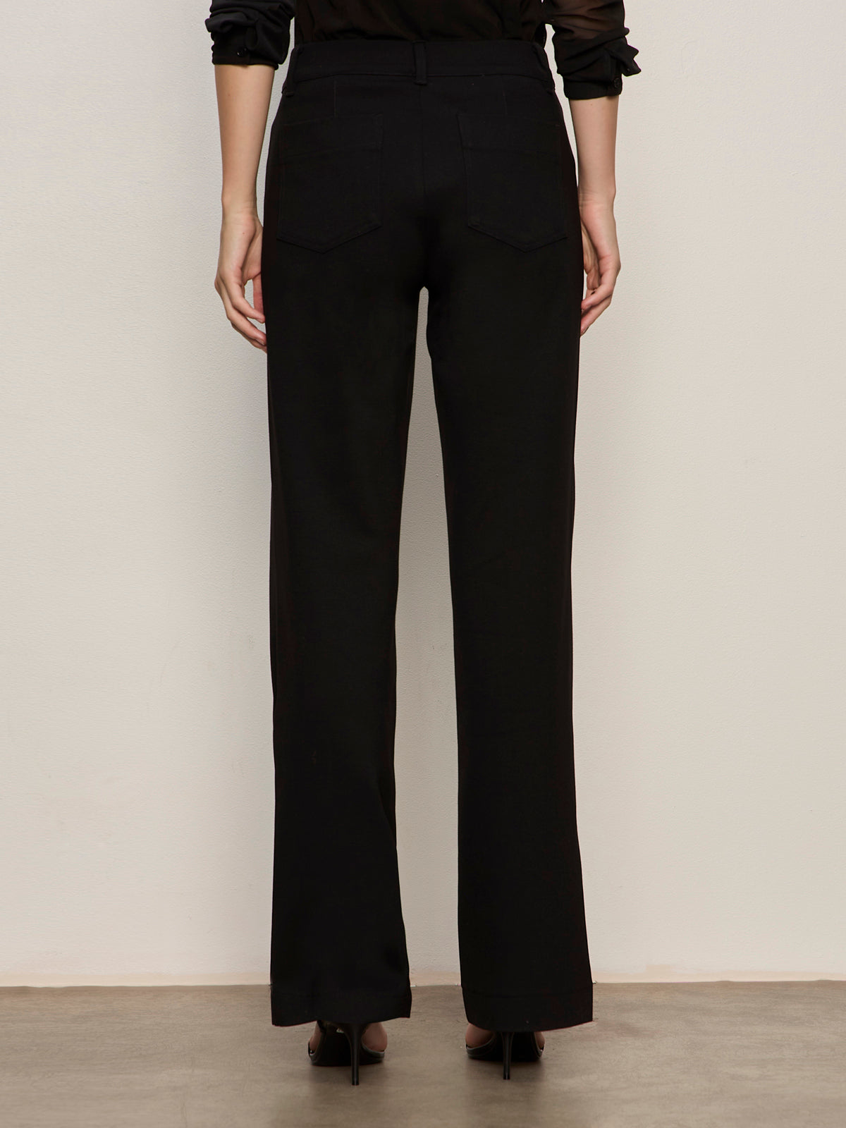 A person stands facing a light wall, wearing the Sanctuary Clothing marine ponte pant in black with black heeled shoes. The photo, taken from behind, highlights the back pockets and straight fit of these high-waisted, wide-leg pants.