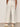 A person’s lower half is shown indoors, wearing Sanctuary Clothing’s lily cropped pant in setting sun stripe—a cream wide-leg pant with thin vertical stripes—paired with white sneakers.