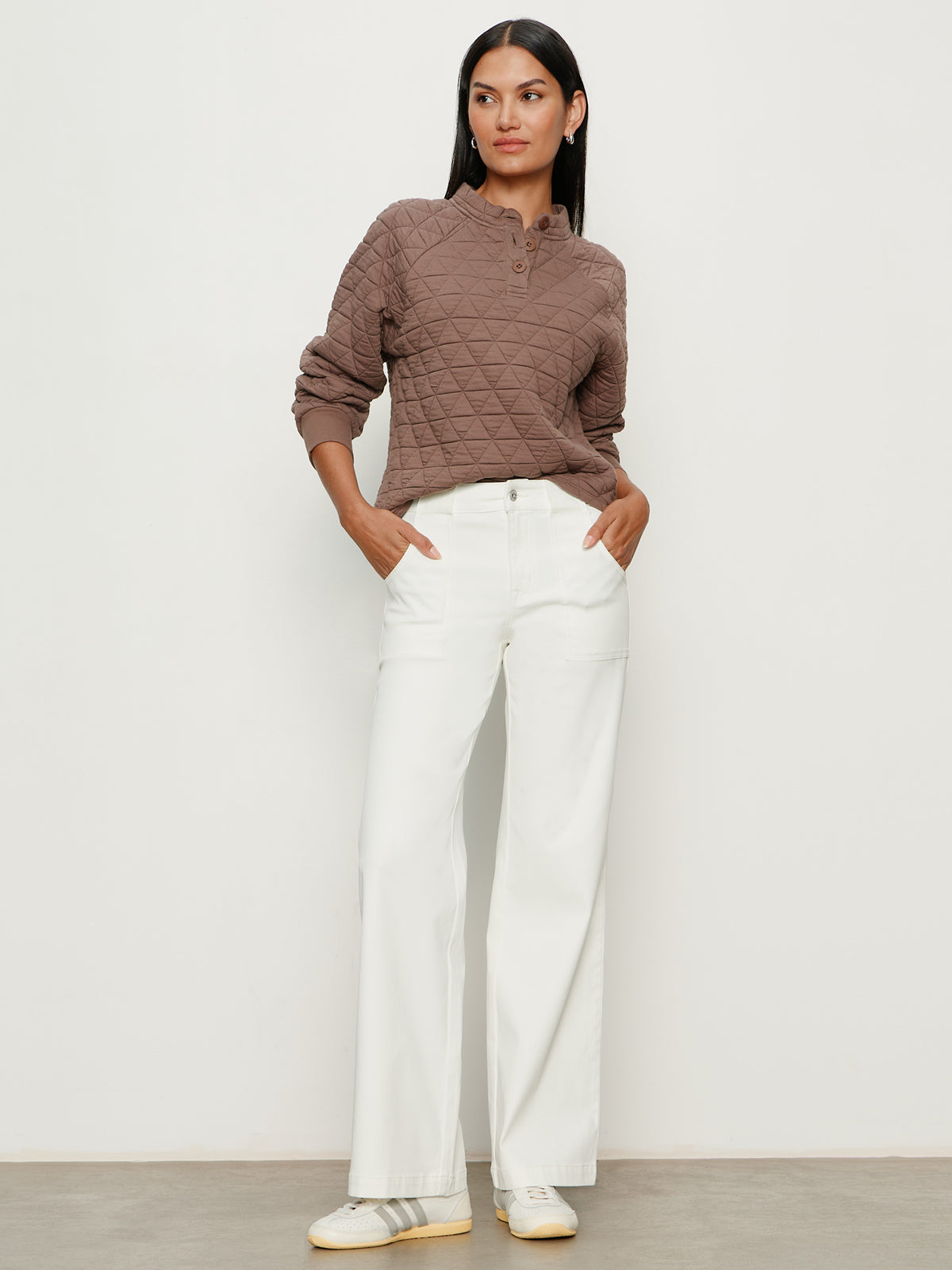 A woman stands against a plain light wall, wearing a textured brown long-sleeve top and Sanctuary Clothing’s sculpted insider wide leg pant in chalk. She has straight dark hair, white sneakers, and her hands in her pockets.
