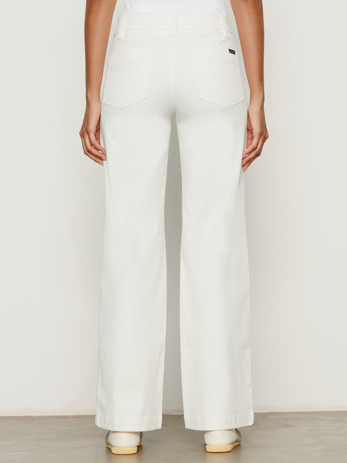 A person stands facing away, wearing Sanctuary Clothing's sculpted insider wide leg pant in chalk with large back pockets and beige shoes, against a plain light background.