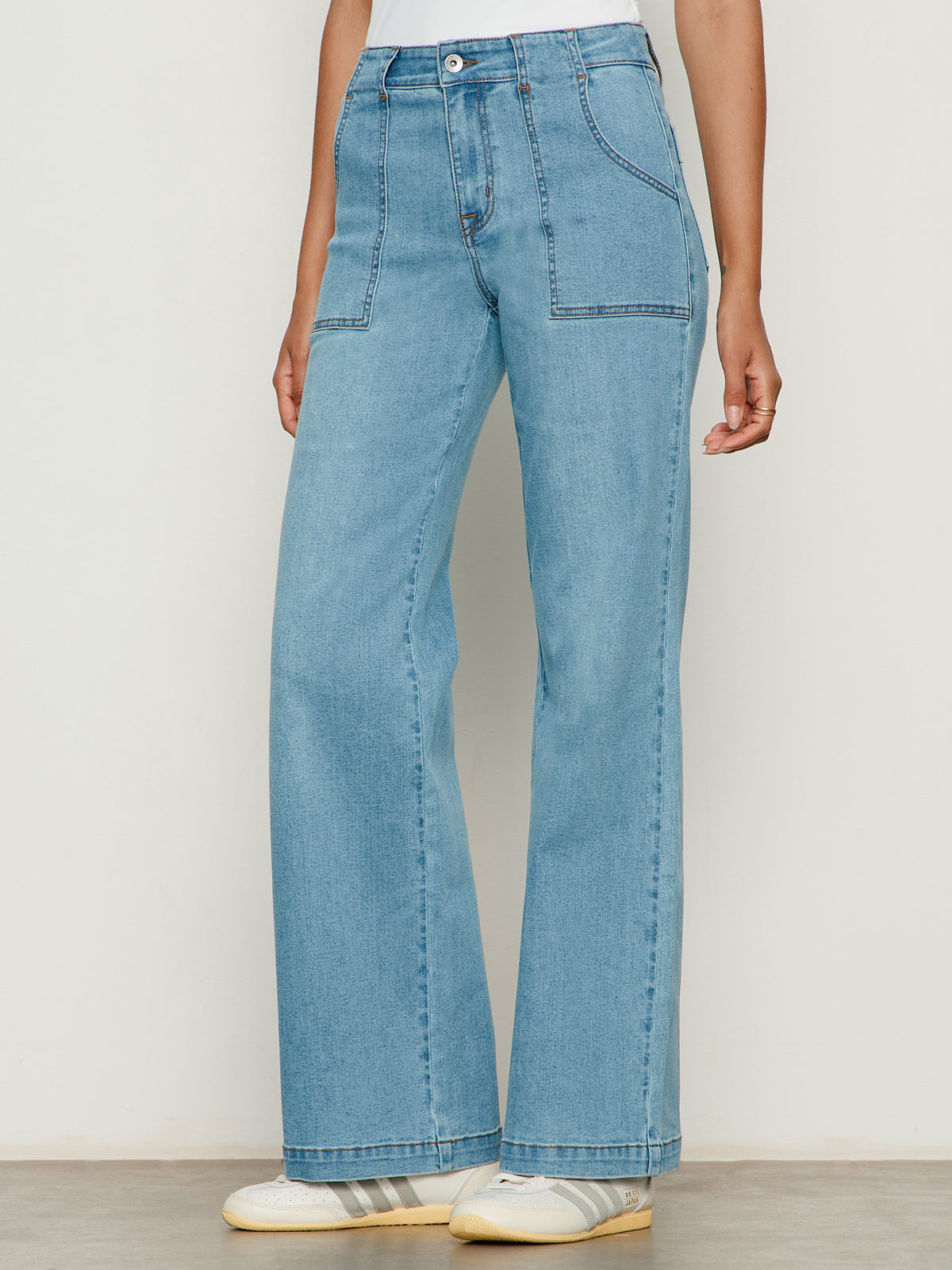 A person is wearing Sanctuary Clothing’s sculpted insider wide leg jean keep up in light blue denim with front patch pockets, paired with white sneakers with beige striped accents, standing against a plain light-colored background.