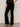 A person stands against a light background, wearing Sanctuary Clothing's velveteen brook bootcut pant in black petites, a black sequin top, and black strappy heeled sandals. Their left hand rests on their hip.