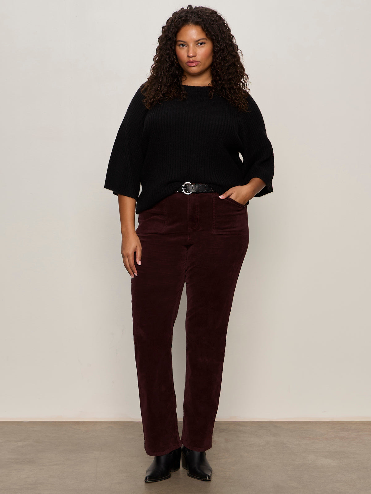 A woman with curly hair stands against a plain light wall, wearing Sanctuary Clothing's velveteen brook bootcut pant in dark crimson (extended sizing), a black knit sweater, and black ankle boots, with one hand in her pocket and a neutral expression.