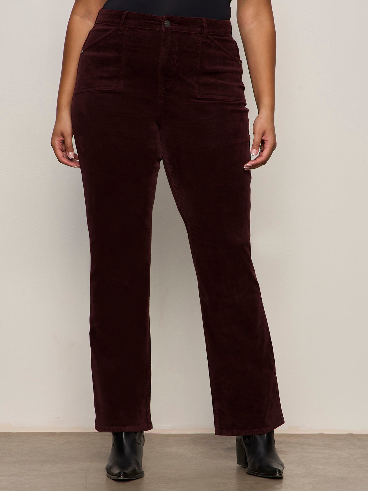 A person stands against a plain light wall, hands relaxed, wearing Sanctuary Clothing's velveteen brook bootcut pant in dark crimson (extended sizing) paired with black boots.