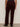 A person is standing indoors facing away, wearing Sanctuary Clothing's velveteen brook bootcut pant in dark crimson (extended sizing) with black heeled shoes against a plain, neutral background.