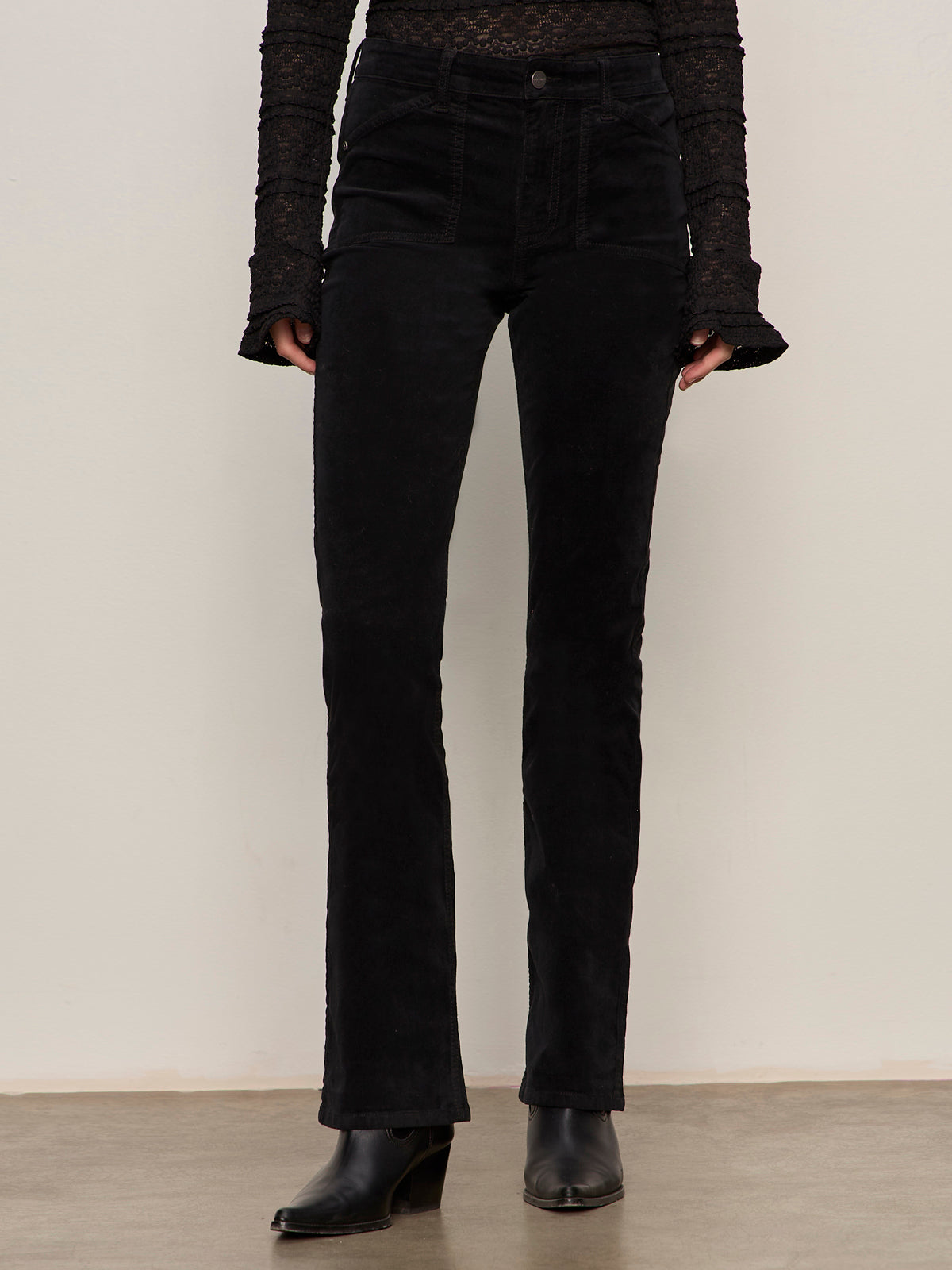 Wearing Sanctuary Clothing's velveteen brook bootcut pant in black with a black, flared-cuff long-sleeve top and black ankle boots, the person stands against a plain light wall, shown from shoulders to feet.