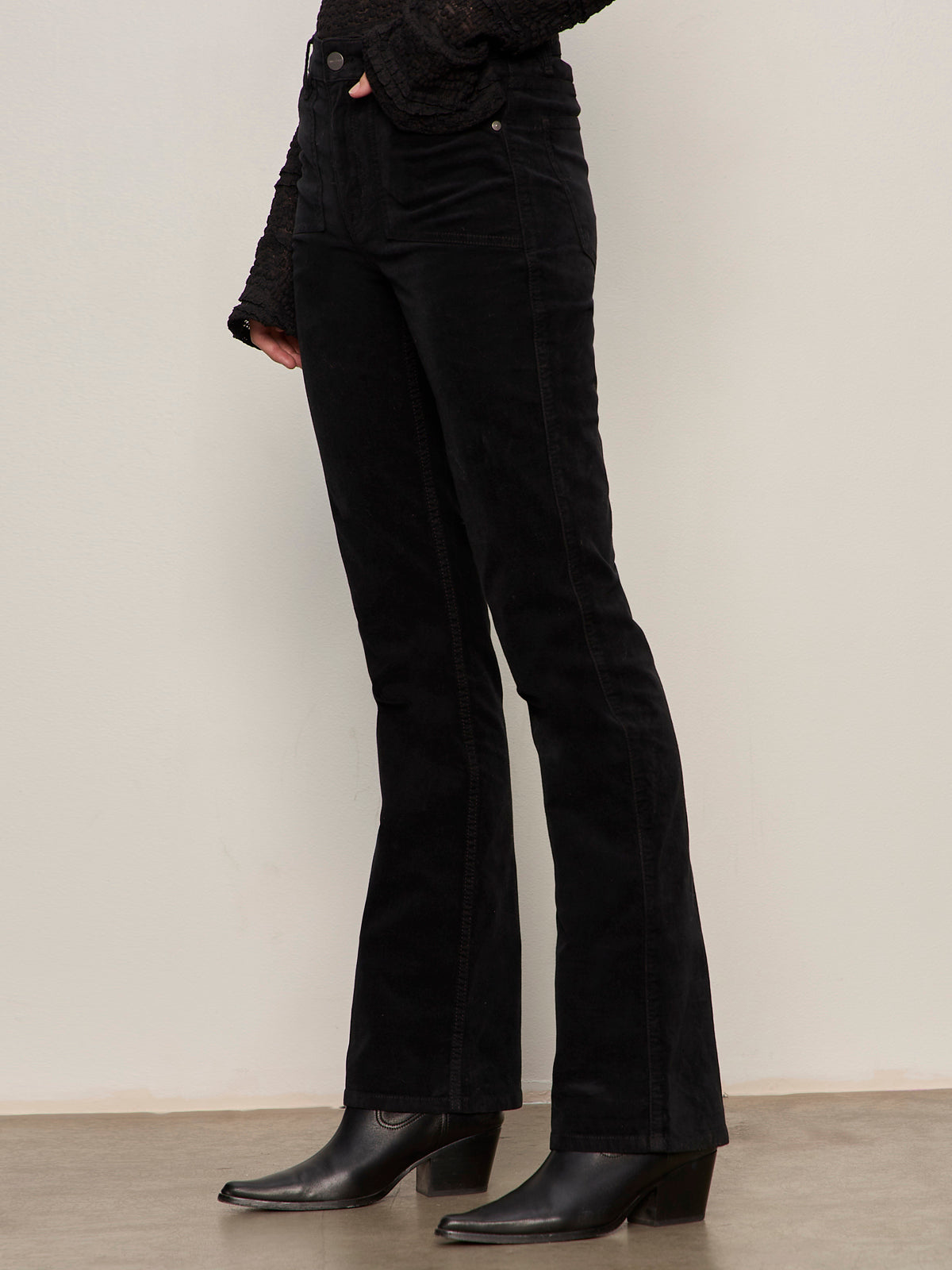 A person wears Sanctuary Clothing's velveteen brook bootcut pant in black with a textured black long-sleeve top and heeled ankle boots, standing against an off-white wall. The photo is cropped above the shoulders.