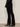 A person wears Sanctuary Clothing's velveteen brook bootcut pant in black with a textured black long-sleeve top and heeled ankle boots, standing against an off-white wall. The photo is cropped above the shoulders.