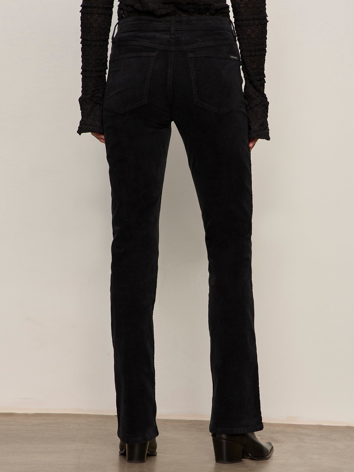 Shown from the back against a plain light background, only the lower body is visible, wearing Sanctuary Clothing velveteen brook bootcut pant in black paired with black heeled shoes.
