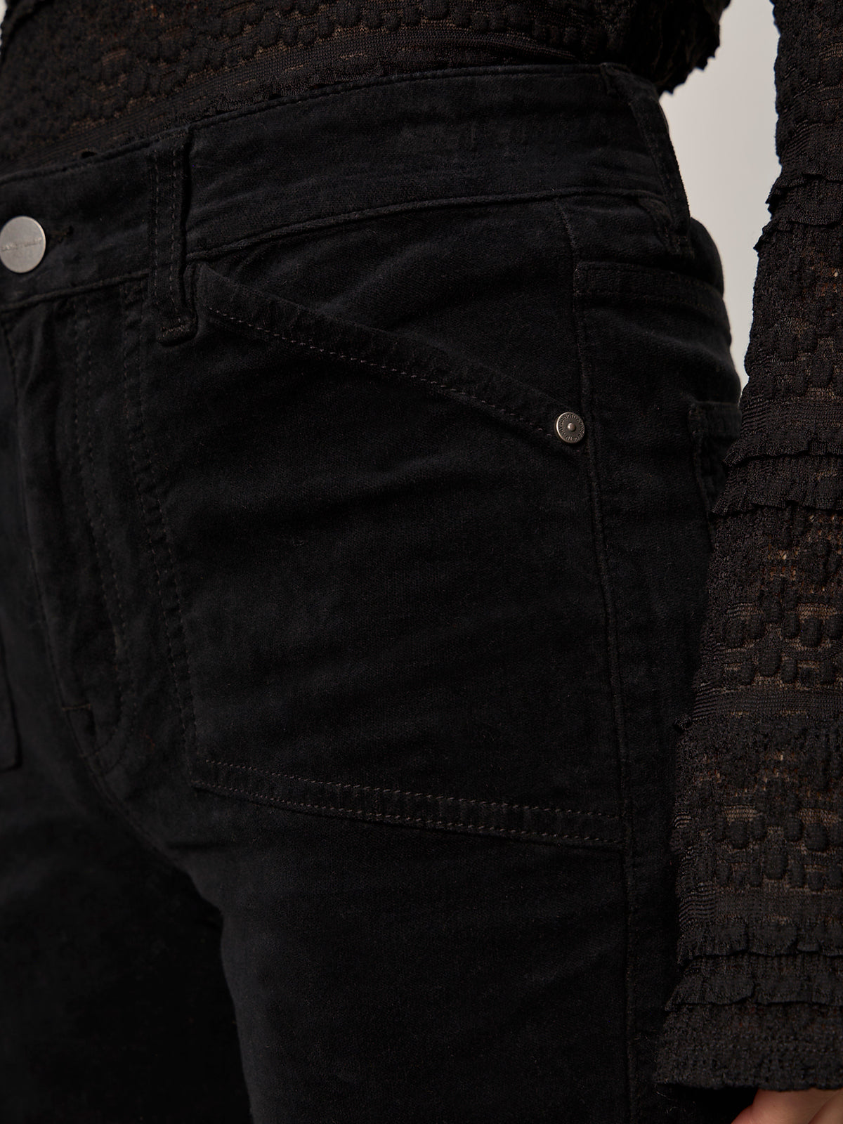 Close-up of a person wearing Sanctuary Clothing's Velveteen Brook Bootcut Pant in black, featuring visible detailed stitching, pocket accents, and paired with a textured, lace-like ruffled long-sleeve top.