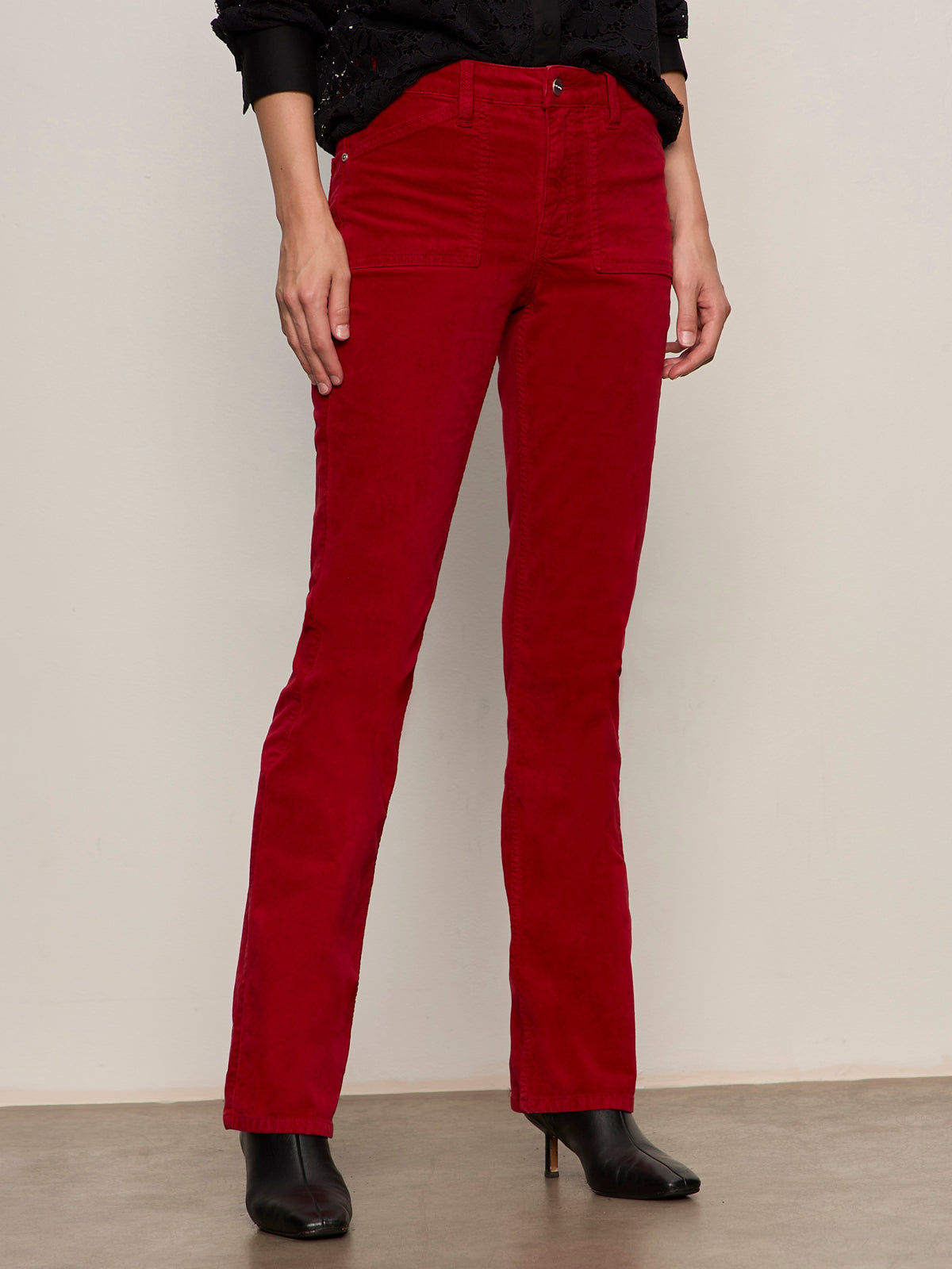 A person wears Sanctuary Clothing's velveteen brook bootcut pant in cherry lacquer (petite), styled with a black long-sleeve top and black heeled ankle boots, standing against a neutral background.