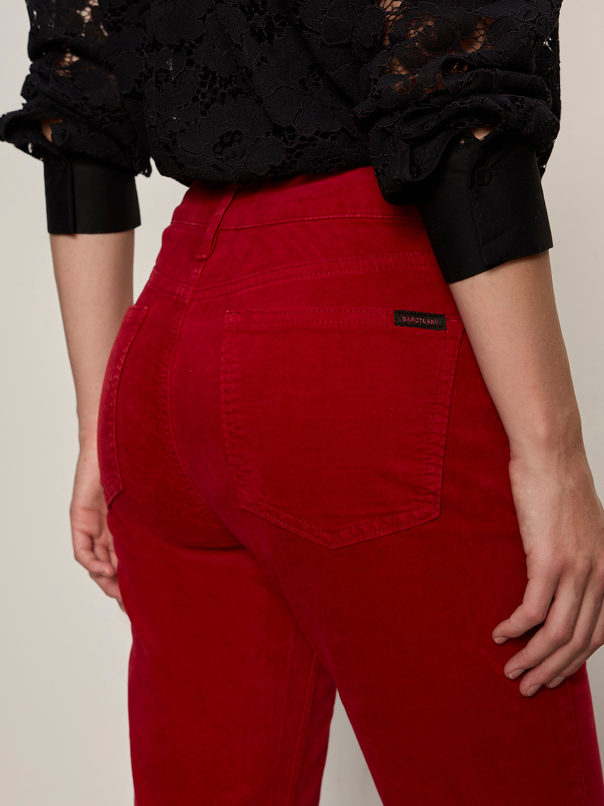 A person wears cherry lacquer velveteen brook bootcut pants by Sanctuary Clothing and a black long-sleeve lace top, with rolled-up sleeves, back pocket, and brand label on display.
