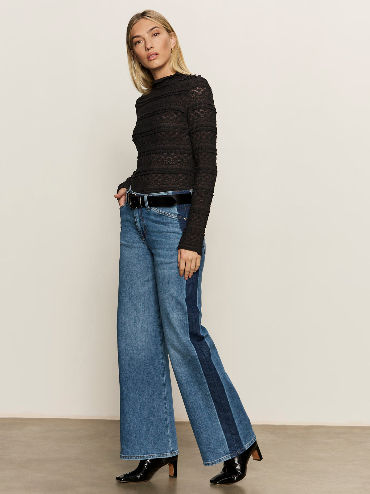 A woman with straight blonde hair and a neutral expression stands against a plain background, wearing Sanctuary Clothing„¢s tuxedo relaxed straight jean flirt with a dark blue side stripe, paired with a black lace top, black belt, and black boots.