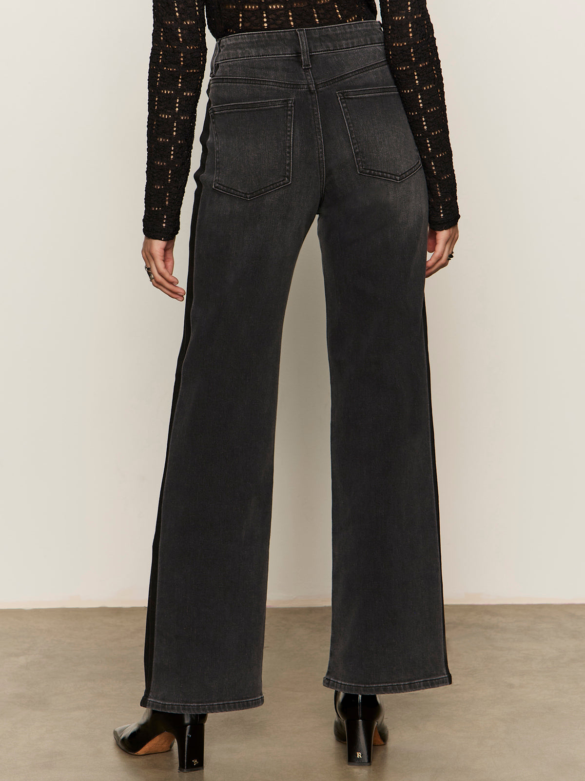 A person faces away, showcasing the back of Sanctuary Clothing's tuxedo relaxed straight jean black tie, paired with a black long-sleeve top and black heeled boots against a plain background.