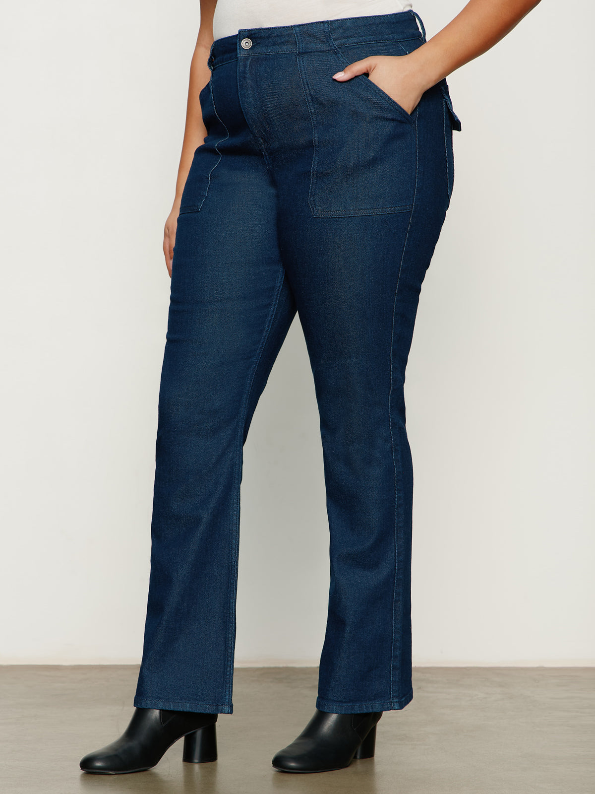 A person is shown from the waist down wearing Sanctuary Clothing's hayden bootcut jean plot twist extended sizing against a white background. The dark blue jeans have front pockets and are styled with black ankle boots.