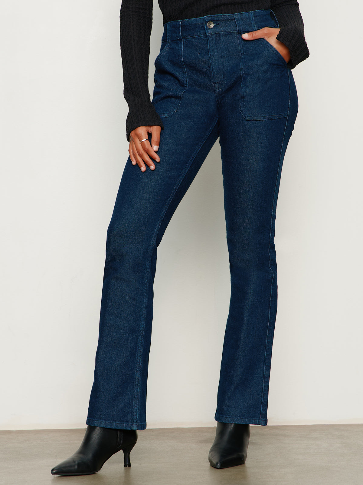A person in Sanctuary Clothing's hayden bootcut jean plot twist, paired with a black ribbed long-sleeve top and black heeled ankle boots, stands against a plain light background with their left hand on their hip.