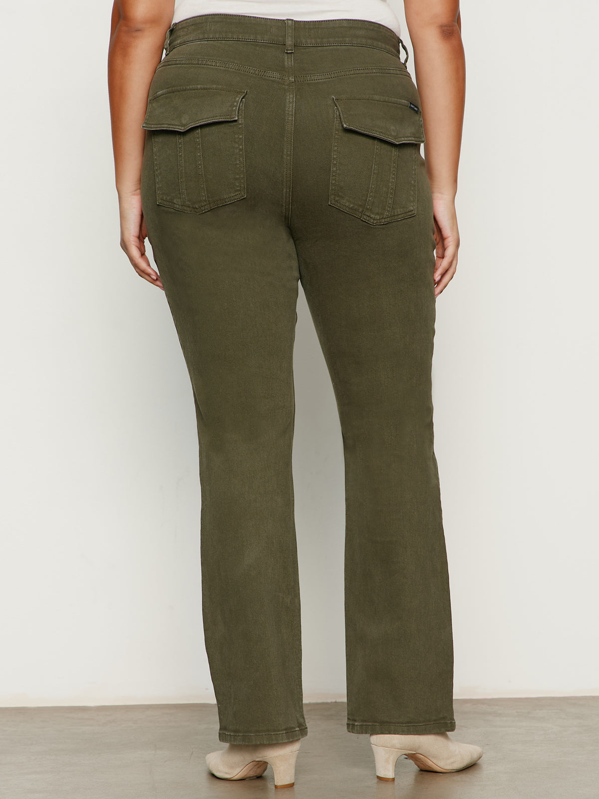 A person is seen from behind wearing Sanctuary Clothing’s hayden bootcut jean in kalamata (olive green) with large back flap pockets and beige heeled shoes, set against a plain light-colored background.