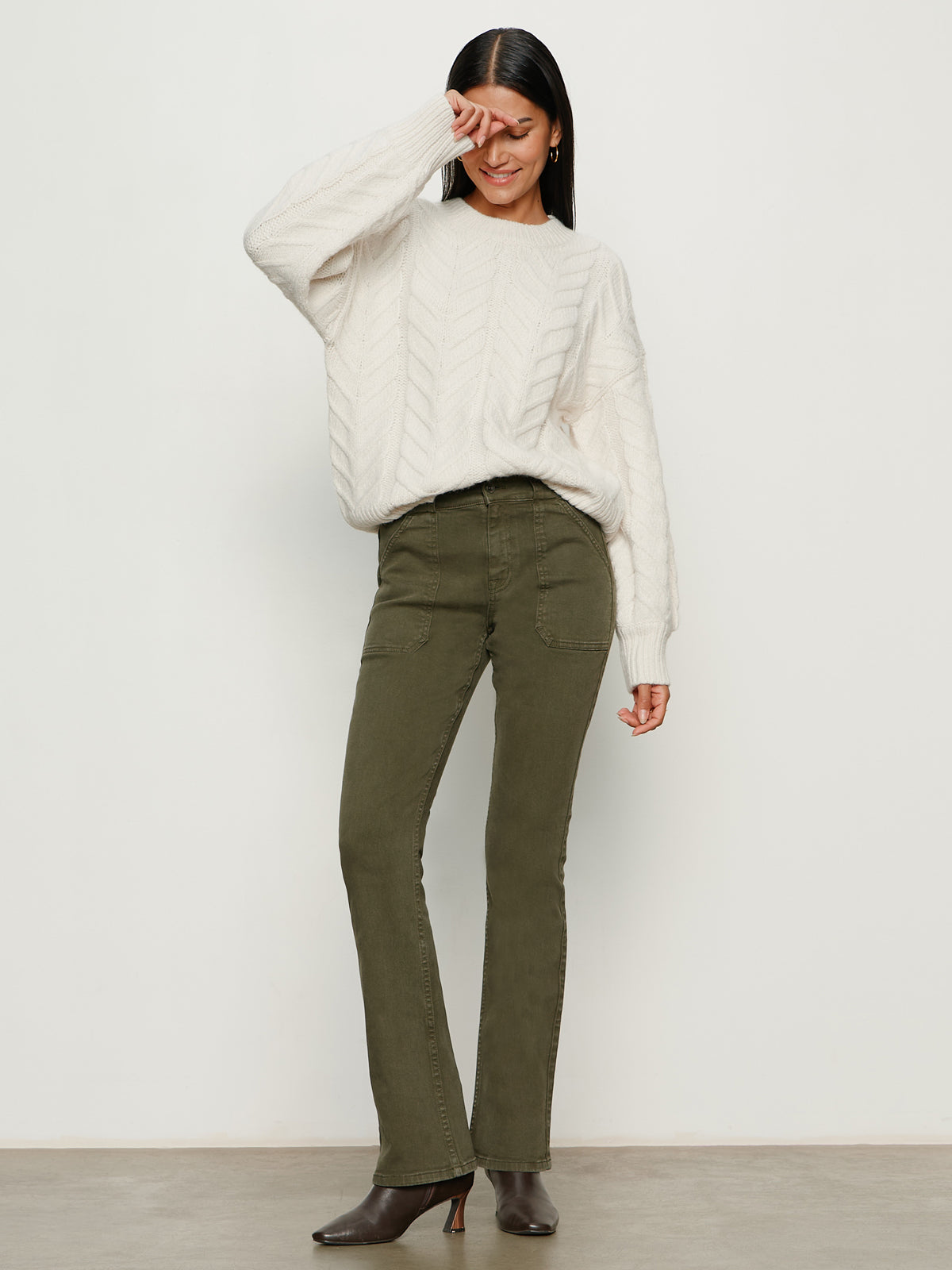 A woman poses against a plain background, smiling playfully as she wears Sanctuary Clothing’s hayden bootcut jean in kalamata with a loose white sweater and brown heeled boots.