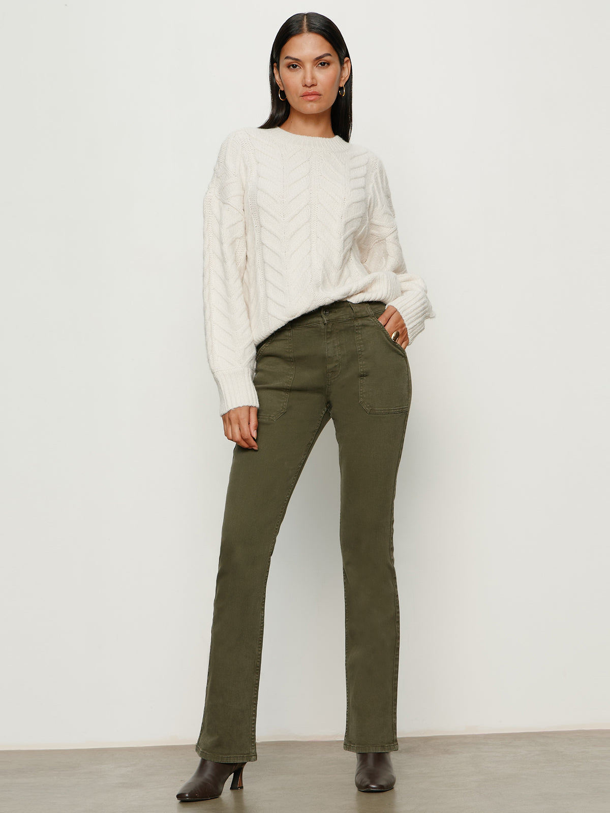 A woman with straight dark hair and a neutral expression stands against a light background, wearing the Sanctuary Clothing hayden bootcut jean in kalamata and a white textured knit sweater, paired with dark heeled ankle boots.