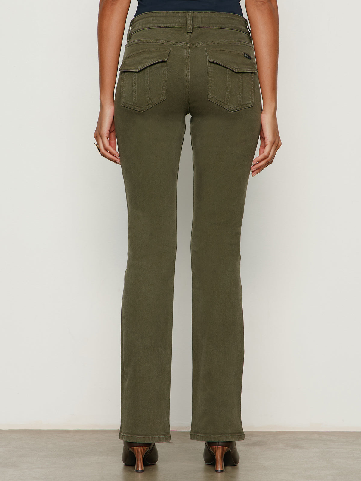 A woman faces away wearing Sanctuary Clothing's hayden bootcut jean in kalamata, featuring high-waisted, olive green flare jeans with flap back pockets, paired with a black top and brown heels.