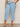 A person wearing Sanctuary Clothing's retreat crop jean skyline extended sizing in light blue, with front patch pockets, styled with a floral top and beige woven sandals, stands on a neutral floor against a plain background.