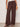 Someone wearing Sanctuary Clothing’s Shoreline Linen Pant in Rich Soil—loose, dark brown drawstring pants—paired with a white sleeveless top, black studded sandals, and chunky bangles, stands against a plain light background.
