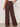 A person stands facing away, wearing Sanctuary Clothing's shoreline linen pant in rich soil. The high-waisted, wide-leg brown pants are styled with a sleeveless top, chunky bangles on one wrist, and set against a plain light background.