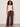 A woman with curly hair poses confidently against a plain background in Sanctuary Clothing’s shoreline linen pant in rich soil, paired with a sleeveless cream henley top, black sandals, and gold bangles.