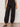 A person wears the Sanctuary Clothing "melody micro linen balloon black" pants, featuring a loose fit and elastic ankle cuffs, paired with a black top and studded sandals, standing against a plain light background.