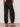 A person stands facing away wearing Sanctuary Clothing's melody micro linen balloon black pants with elastic cuffs, paired with a black top and studded sandals on a neutral floor against a plain light wall.