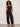 A woman with curly hair and sunglasses poses against a light background, wearing Sanctuary Clothing's melody micro linen balloon black pants, a black sleeveless top, statement necklace, and black sandals with one hand in her pocket.