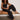 A woman with curly hair, sunglasses, and studded sandals sits casually on a tan bench wearing the Melody Micro Linen Balloon Black tank top by Sanctuary Clothing and black jogger pants against a plain wall.
