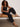 A woman with curly hair, sunglasses, and studded sandals sits casually on a tan bench wearing the Melody Micro Linen Balloon Black tank top by Sanctuary Clothing and black jogger pants against a plain wall.
