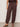 The lower half of a person is shown wearing Sanctuary Clothing’s melody micro linen balloon rich soil pants—loose brown drawstring bottoms with elastic cuffs—paired with a matching top and black studded sandals, against a plain light wall.