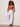 A woman with curly hair and sunglasses stands against a light wall with hands in her pockets, wearing Sanctuary Clothing's melody micro linen balloon white pants, sandals, and a sleeveless rust-colored top.