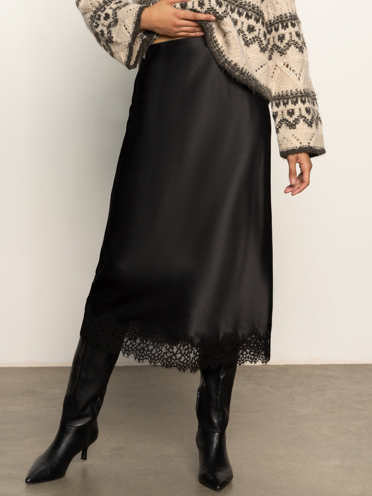Person wearing a black satin skirt with lace hem and black boots against a plain background