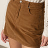 A person wears the Sanctuary Clothing briar corduroy mini skirt in bronze, featuring front pockets and belt loops, paired with a cream knitted sweater. Shown from the waist to mid-thigh.