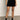 A person wears a white top, black socks, black heeled shoes, and the quilted velvet mini skirt in black by Sanctuary Clothing while standing indoors on a concrete floor against a plain light-colored wall.
