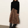 A woman stands against a plain wall wearing Sanctuary Clothing's nostalgia pleated midi skirt in mocha leo, paired with a black sweater and knee-high black boots. She looks down with her hands by her sides.