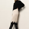 A woman with straight light brown hair and a neutral expression stands against a white wall wearing Sanctuary Clothing's evermore pleated midi skirt in light oat, styled with a black turtleneck sweater and black knee-high heeled boots.
