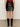 A person stands indoors, shown from the back, wearing the Sanctuary Clothing broadway leather skirt in black and matching black knee-high boots. The floor is gray and the wall is off-white.
