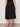 A person stands on a concrete floor wearing the Sanctuary Clothing "matter of time poplin skirt" in black, paired with a brown bracelet and black sandals. Their back is visible from shoulders down, set against a plain light-colored wall.